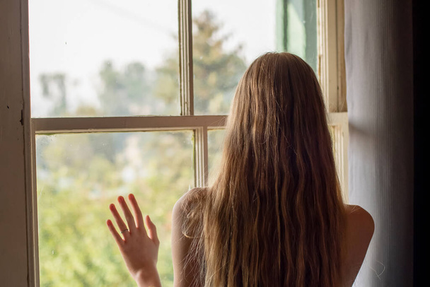 Evde kal: odanın içindeki puslu pencerenin yanındaki yalnız genç kız. Kişisel tecrit koronavirüs konsepti - Fotoğraf, Görsel