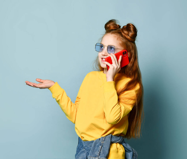 Teenager red-haired girl in blue smoked glasses, yellow sweatshirt and glossy black skirt is discussing news by phone - Foto, afbeelding