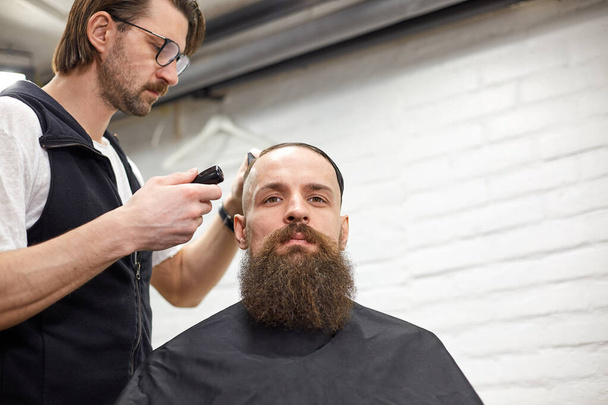 現代の理髪店で残忍な男。美容師は長いひげを生やした男の髪型になります。美容師ははさみや櫛でヘアスタイルを行います - 写真・画像
