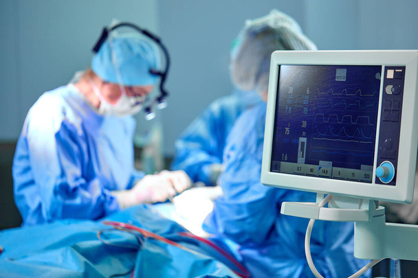 Ηλεκτροκαρδιογράφημα στο νοσοκομείο χειρουργική επέμβαση στα επείγοντα περιστατικά δείχνει καρδιακό ρυθμό του ασθενούς με θολή ομάδα χειρουργών φόντο - Φωτογραφία, εικόνα