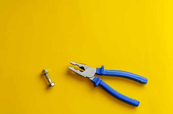 Pliers and bolt on a yellow background. Composition of hand tools with blue handles and one bolt with nut. View from above. Selective focus. - Photo, image