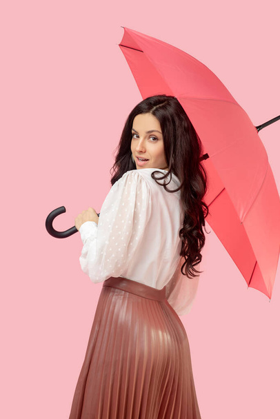 Young pretty dark-haired woman in a pleated skirt holding a red umbrella and looking flirty - Fotoğraf, Görsel