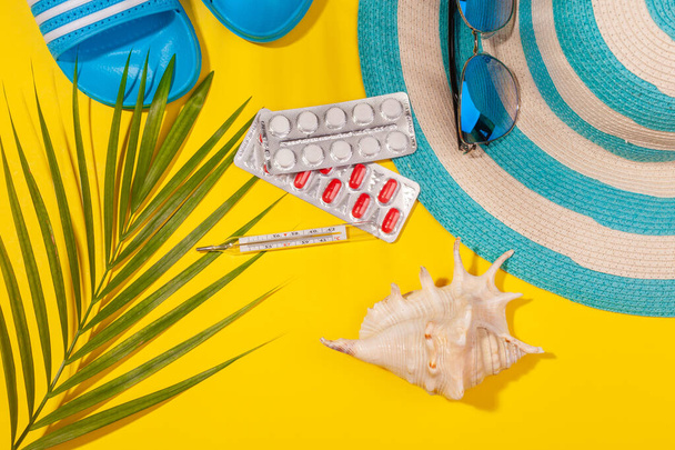 Top view on white and red pills in blister pack, thermometer, blue hat, blue flip-flops, sunglasses, seashell, palm leaf on yellow background. Concept of beach holiday, sea tour, warm sunny summer - Φωτογραφία, εικόνα
