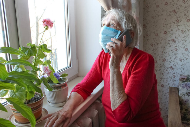 Koncepcja Covid-19, zdrowie, bezpieczeństwo i pandemia - starsza samotna kobieta nosząca ochronną maskę medyczną siedząca w pobliżu okna w domu dla ochrony przed wirusem - Zdjęcie, obraz