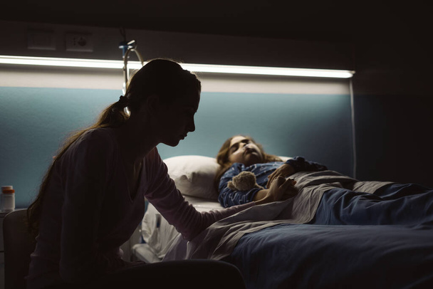 Zoufalá mladá matka pomáhá své nemocné dítě ležící v nemocniční posteli v noci - Fotografie, Obrázek