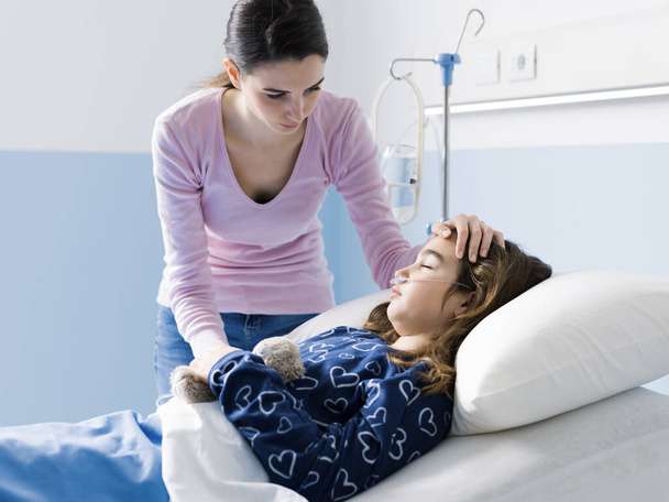 Young woman comforting her sick child lying in bed at the hospital, she is touching her head, pediatrics and healthcare concept - Foto, immagini