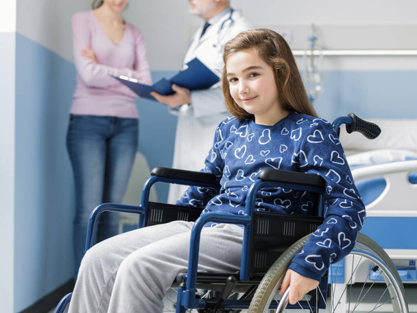 Улыбающаяся девушка в инвалидном кресле в больнице, ее мать и врач разговаривают на заднем плане
 - Фото, изображение