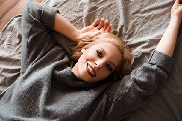 Beautiful young woman in gray sweater laughing lying in bed - Fotoğraf, Görsel