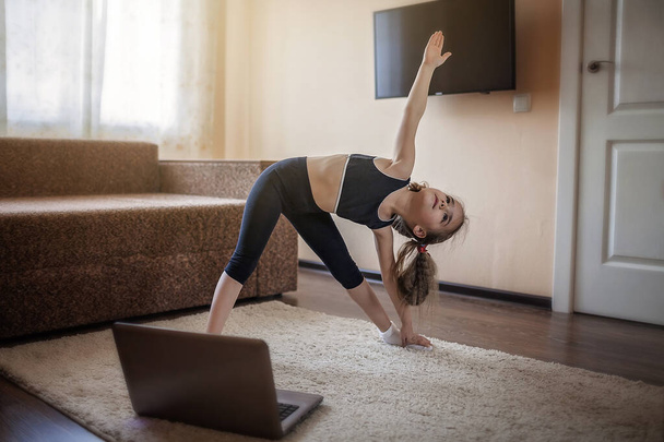 Όμορφη νεαρή κοπέλα στα αθλητικά βλέποντας online βίντεο στο laptop και κάνοντας ασκήσεις γυμναστικής στο σπίτι. Μακρινή εκπαίδευση με προσωπικό γυμναστή, κοινωνική απόσταση ή αυτο-απομόνωση, έννοια της online εκπαίδευσης - Φωτογραφία, εικόνα