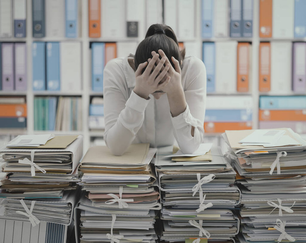 Frustrated you office worker leaning on piles of paperwork, she is overloaded with work and stressed - Photo, Image