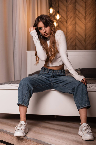 Модная сексуальная молодая женщина-модель в модном белом топе в синих джинсах в модных кроссовках сидит на винтажной кровати в спальне. Стильная красивая девушка отдыхает в уютной студии
. - Фото, изображение