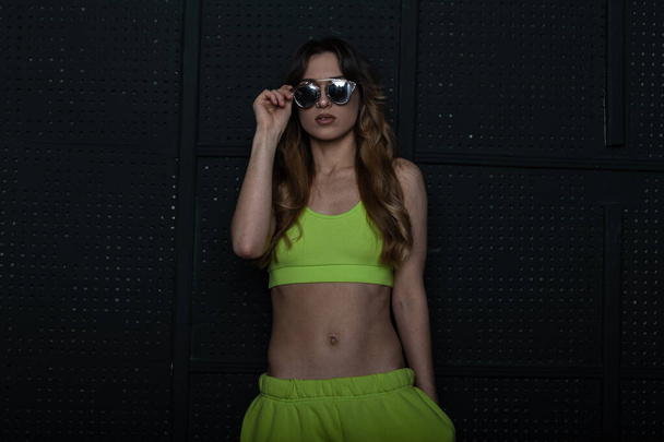 Pretty cool young woman hipster in stylish sportswear straightens vintage sunglasses indoors. Attractive girl model in a trendy light green tracksuit posing near a vintage black wall in the room. - Φωτογραφία, εικόνα