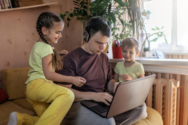 Gelukkige man met vrolijke kinderen die laptop en oortelefoon gebruiken terwijl hij thuis op de bank zit, thuiskantoor met kinderen, leven tijdens quarantaine - Foto, afbeelding