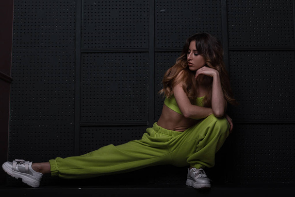 Fashionable beautiful slim model of a young woman in a stylish light green sports suit is engaged in fitness near the vintage black wall in the room. Attractive girl doing stretching in the studio - Valokuva, kuva