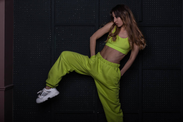Model of a young beautiful woman dancer in a fashionable green suit in stylish white sneakers ποζάρουν κοντά σε vintage μαύρο τοίχο σε ένα δωμάτιο. Κοκκινομάλλα μοντέρνα κοπέλα ποζάρουν στο δωμάτιο. - Φωτογραφία, εικόνα
