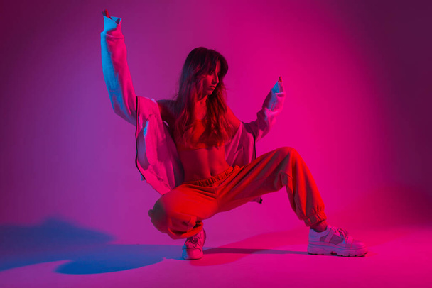 Glamour young woman dancer in fashionable youth clothes in a white sneakers sits in a room with bright pink light. Modern girl model poses in the studio with neon amazing colorful red color. - Photo, Image