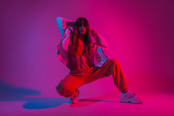 Αμερικανίδα νεαρή χορεύτρια με μοντέρνα νεανικά ρούχα σε λευκό sneakers κάθεται σε ένα δωμάτιο με έντονο ροζ φως. Αθλητικό όμορφο κορίτσι μοντέλο ποζάρει στο στούντιο με νέον πολύχρωμο φως. - Φωτογραφία, εικόνα