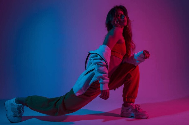 Beautiful young woman dancer is engaged in fitness before dancing. Attractive girl in stylish sportswear posing in the studio with bright blue-pink light. Lady and neon. - Φωτογραφία, εικόνα
