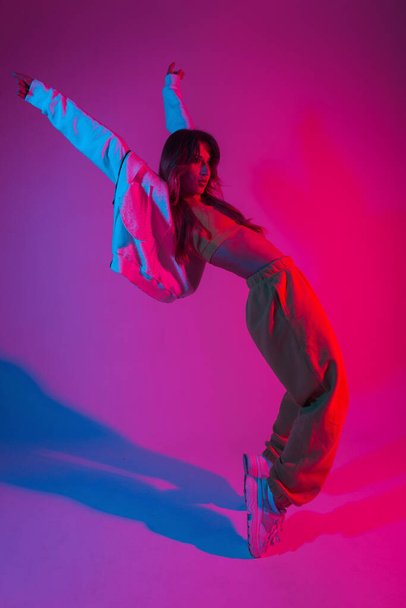 Dançarina muito jovem em roupas jovens na moda em um tênis branco dançando em uma sala com luz vermelha. Moderno modelo de moda linda menina goza de uma dança no estúdio com neon cor rosa-azul
. - Foto, Imagem