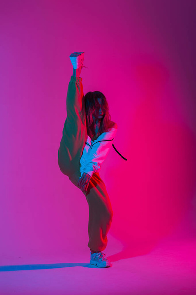 Fashion model young modern woman in stylish clothes in trendy gym shoes dancing indoors with bright multi-colored color. Sporty girl dancer posing in a room with bright neon glamour pink-blue light. - Фото, зображення