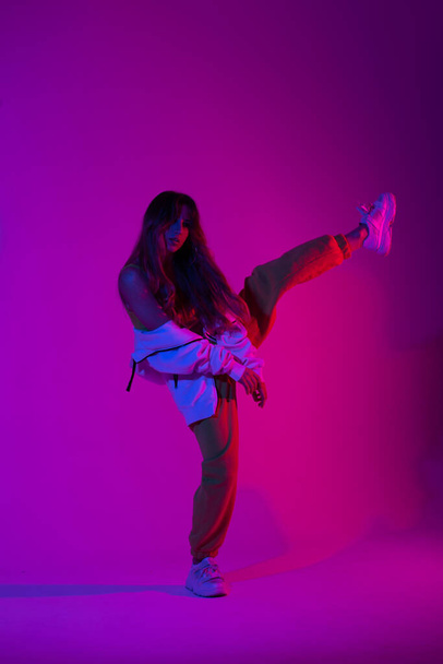 Moderna e elegante jovem dançarina com roupas jovens de esportes da moda em tênis de moda fica em uma perna no estúdio com uma luz azul brilhante. Menina glamour dançando no quarto com cor de néon rosa
. - Foto, Imagem