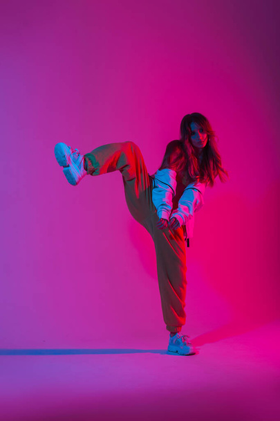 Stylish young woman dancer with trendy sports youth clothes in fashion sneakers stands on one leg in the studio with a bright pink light. Modern girl dancing in the room with multi-colored neon color. - 写真・画像