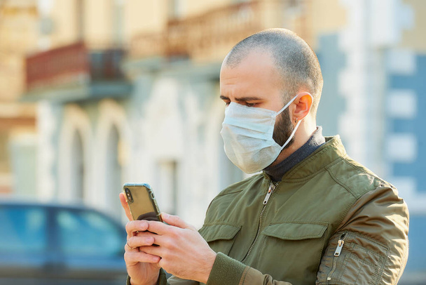 Un homme caucasien portant un masque médical pour éviter la propagation du coronavirus (COVID-19). Un mec avec un masque chirurgical sur le visage à l'aide d'un smartphone dans une rue
. - Photo, image