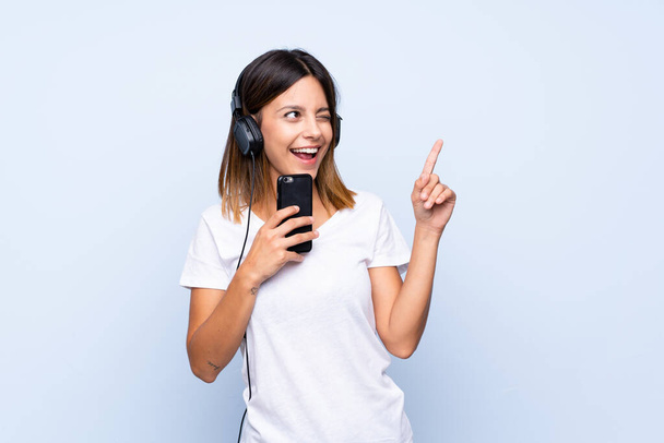 Jonge vrouw over geïsoleerde blauwe achtergrond met behulp van de mobiele telefoon met koptelefoon en zingen - Foto, afbeelding