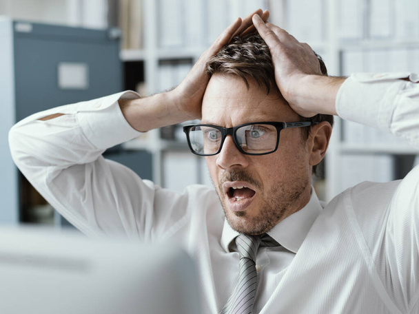 Panicking businessman with head in hands in his office, he is receiving shocking bad news on his computer - 写真・画像