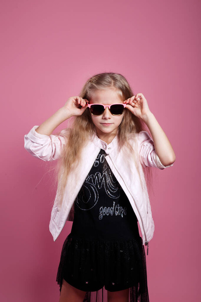 fashionable little girl wearing black dress, sunglasses and pink leather jacket posing on pink background - Foto, imagen