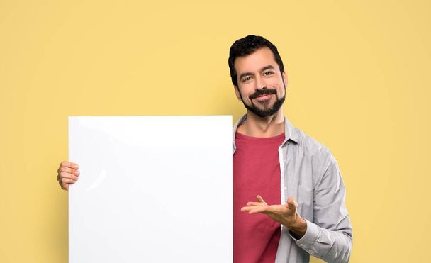 Handsome man with beard holding an empty placard over isolated yellow background - Fotoğraf, Görsel