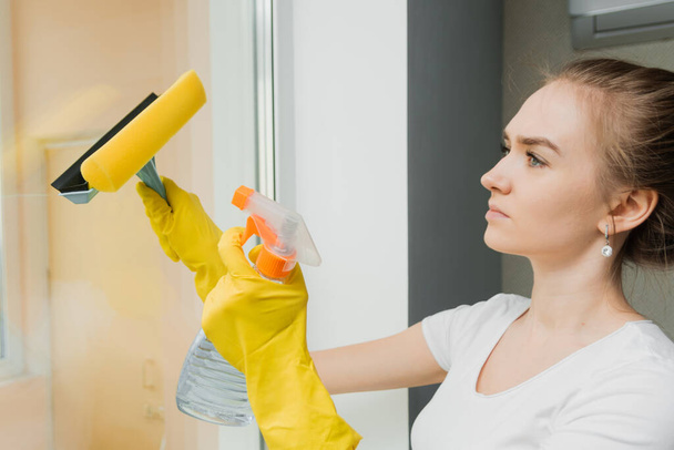 Cute adult girl washes a window with a scraper. Spring cleaning. An employee of a cleaning company wipes plastic windows in the house. - Photo, Image