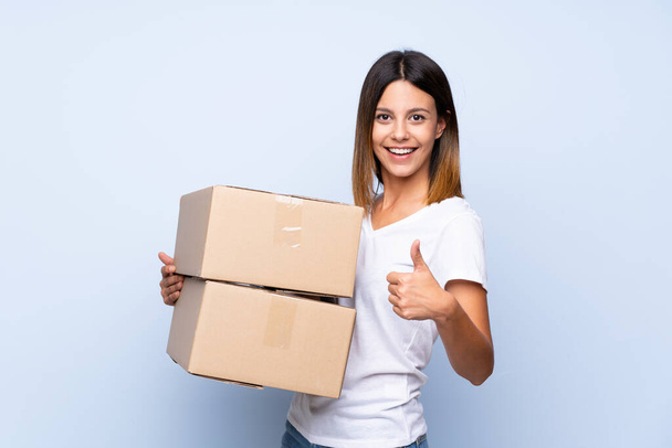 Young woman over isolated blue background holding a box to move it to another site with thumb up - Photo, image