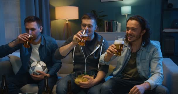 Three handsome Caucasian friendly men doing cheers with beer and cheering up for football game on TV while sitting on sofa at night. Male friends watching sport channel late in evening. - Video