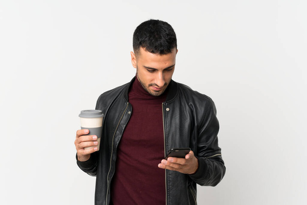 Young man over isolated white background holding coffee to take away and a mobile - Фото, зображення