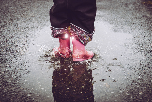 Ragazza divertendosi, saltando in pozzanghera d'acqua sulla strada bagnata, indossando stivali da pioggia con strisce di tessuto riflettente dettaglio brillante. Elevata visibilità e sicurezza nell'oscurità
. - Foto, immagini