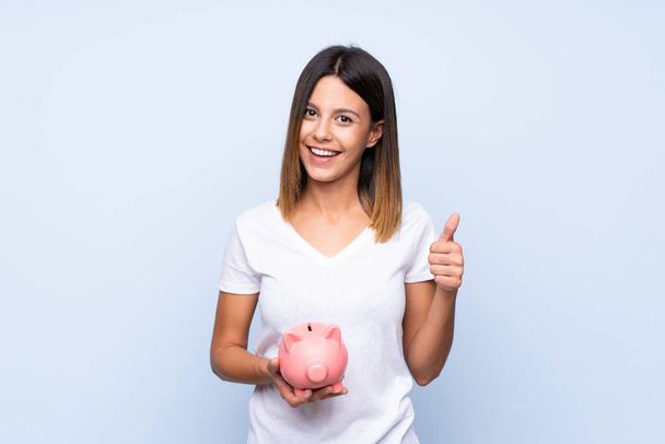 Young woman over isolated blue background holding a big piggybank - Fotografie, Obrázek