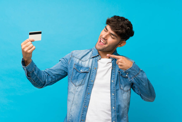 Молодой красивый мужчина на изолированном синем фоне держит кредитку
 - Фото, изображение