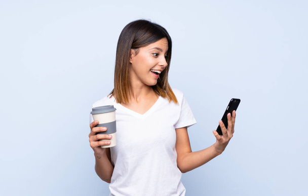 Young woman over isolated blue background holding coffee to take away and a mobile - Фото, зображення