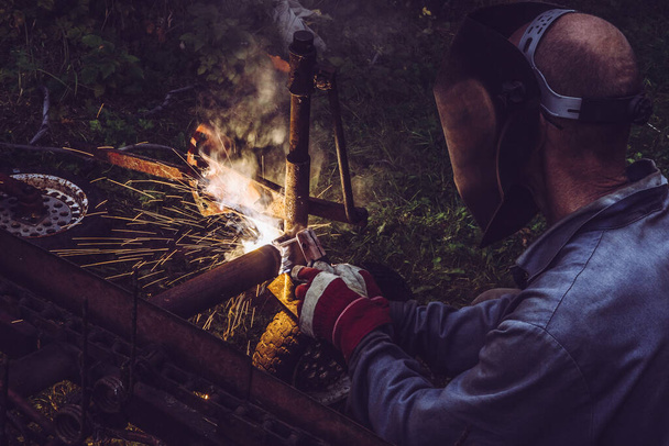 Homem usando máquina de solda vara para cortar ferro fundido construção de metal ao ar livre, usando máscara protetora e luvas, faíscas voar
. - Foto, Imagem