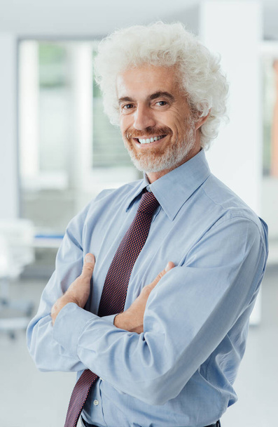 Smiling successful businessman posing with arms folded in his office and smiling at camera, room interior on background - Foto, imagen