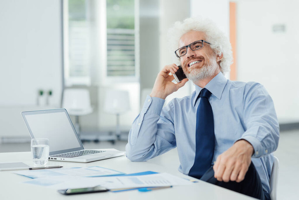 Smiling businessman on the phone sitting at office desk and talking with a customer, consultancy and customer service concept - Фото, изображение