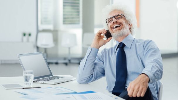 Happy businessman sitting at office desk and laughing on the phone during a break, office interior on background - Photo, image