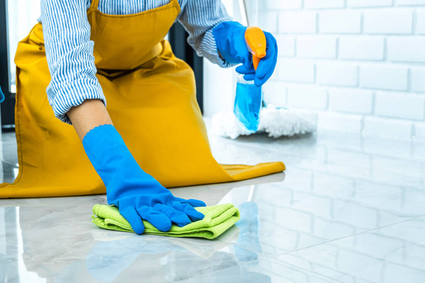 Conceito de limpeza e limpeza da esposa, Jovem feliz em luvas de borracha azul limpando poeira usando um spray e um espanador ao limpar no chão em casa
. - Foto, Imagem