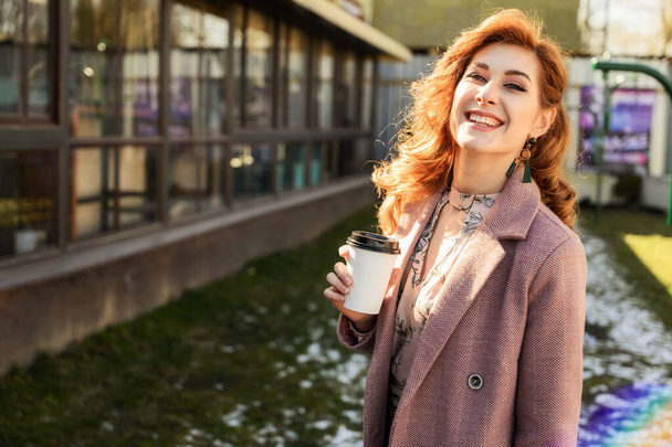 Foto de una hermosa mujer de moda con el pelo largo en la ciudad de primavera bajo el sol al aire libre con una taza de café en las manos
 - Foto, Imagen