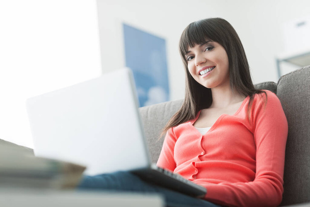Young attractive woman relaxing at home, she is sitting on the couch and connecting to the internet using a laptop - Photo, image