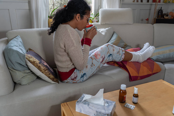 Femme ayant un rhume, assise sur un canapé blanc dans le salon tout en buvant du thé. Jambes en chaussettes chaudes blanches, médicaments et papier de soie sur la table. Concept
. - Photo, image