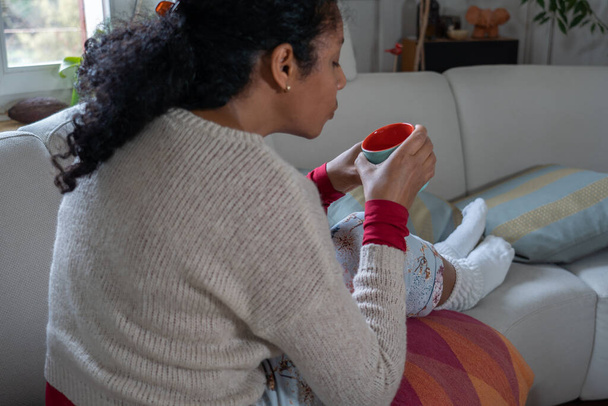 Donna che ha il raffreddore, seduta sul divano bianco in soggiorno mentre beve il tè. Gambe in calzini bianchi caldi, medicina e carta velina in tavola. Concetto
. - Foto, immagini