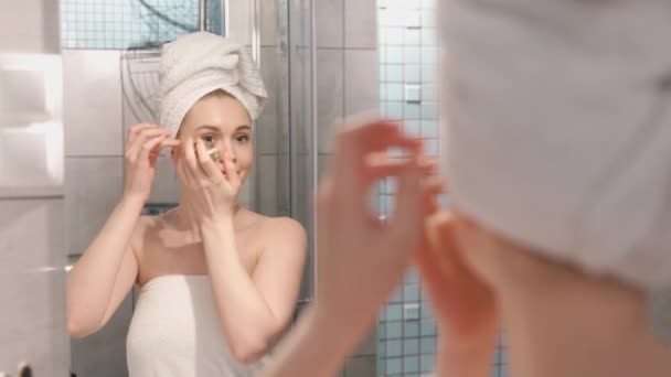 Πορτρέτο φωτογραφία της όμορφης νεαρής γυναίκας με ρόμπα ντους και πετσέτα στέκεται στο μπάνιο και χτυπώντας ενυδατικά μπαλώματα κάτω από τα μάτια της - Πλάνα, βίντεο