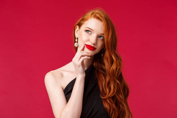 Close-up portrait of dreamy elegant and stylish redhead woman in black dress, red lipstick, making plan, thinking and smiling pleased of finding great idea, touch lip and look away pondering - Foto, imagen